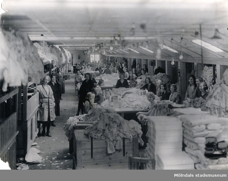 Kvinnliga fabriksarbetare står uppställda för fotografering i fabrikslokal på "Strumpan" (Viktor Samuelsons fabrik), okänt årtal.